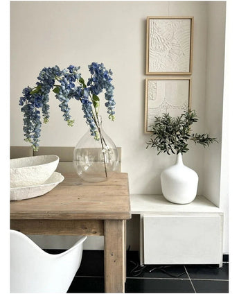 Op een houten tafel staat een doorzichtige vaas met daarin de Kunst Blauweregen van 115 cm, naast een witte schaal. Vlakbij staat een witte vaas met groen op een witte standaard, onder twee ingelijste kunstwerken met een structuur op een lichte muur.