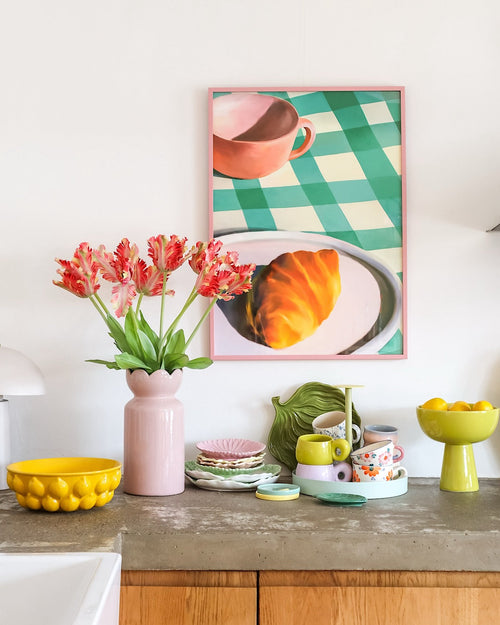 Un piano di lavoro con tutti i tipi di stoviglie colorate e un fiore artificiale Tulipano Parrocchetto francese di 76 cm in rosso/giallo in un vaso rosa. Sopra è appesa la foto incorniciata di un croissant e una tazza da tè su una tovaglia a quadri verdi.