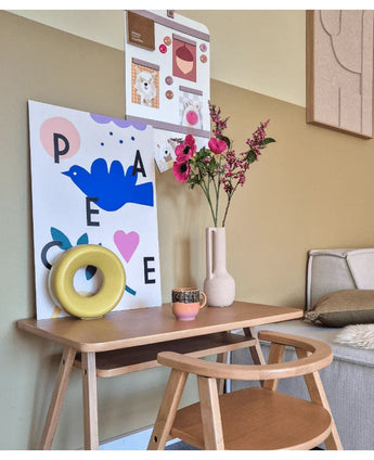 Una piccola scrivania in legno con una sedia è decorata con un fiore artificiale di erba gatta rosa da 75 cm, una tazza, una scultura ad anello gialla e stampe artistiche colorate sulla parete sopra. Sulla destra è parzialmente visibile una panchina.