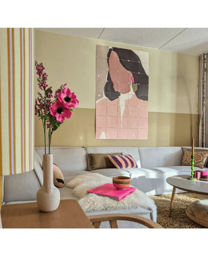Un soggiorno moderno con un divano modulare, una composizione floreale rosa di erba gatta artificiale (75 cm) in un vaso, un soffice plaid e un dipinto murale astratto di una donna. L'interno è caratterizzato da colori tenui e accenti fantasia.