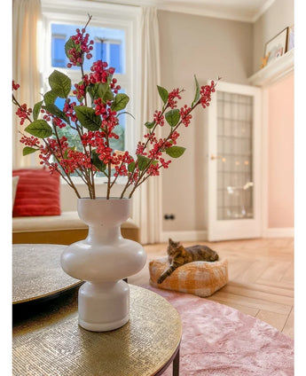 Su un tavolo c'è un vaso bianco con bacche rosa Kunsttak (64 cm) e foglie verdi. Sullo sfondo, un gatto giace su un cuscino rosa in un luminoso soggiorno con pareti beige e una porta a vetri.