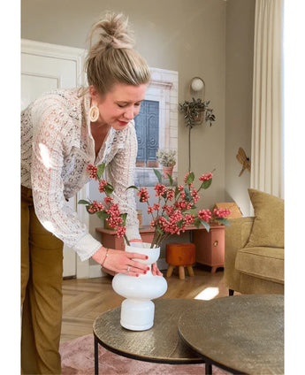 Una donna con i capelli biondi raccolti in uno chignon dispone il Kunsttak Berries rosa da 64 cm in un vaso bianco su un tavolo rotondo in un accogliente soggiorno dai toni della terra e dall'arredamento moderno.