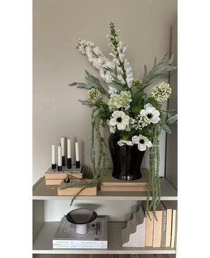 Su uno scaffale contro una parete chiara c'è un vaso nero con il fiore artificiale Amaranthus 124 cm verde, accanto a cinque candele bianche, libri beige e oggetti decorativi dai toni neutri.