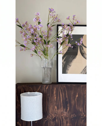 In un vaso trasparente, i fiori della Kunsttak Daphne Spray 108 cm viola sono posizionati su una superficie di legno scuro accanto a un'opera d'arte moderna e astratta in una cornice, con un paralume strutturato bianco sotto.