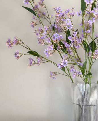 Raffinato Ramo Artificiale di Dafne di 108 cm di colore viola con foglie verdi, disposto in un vaso di vetro trasparente costolato e posto su sfondo a tinta unita di colore chiaro.