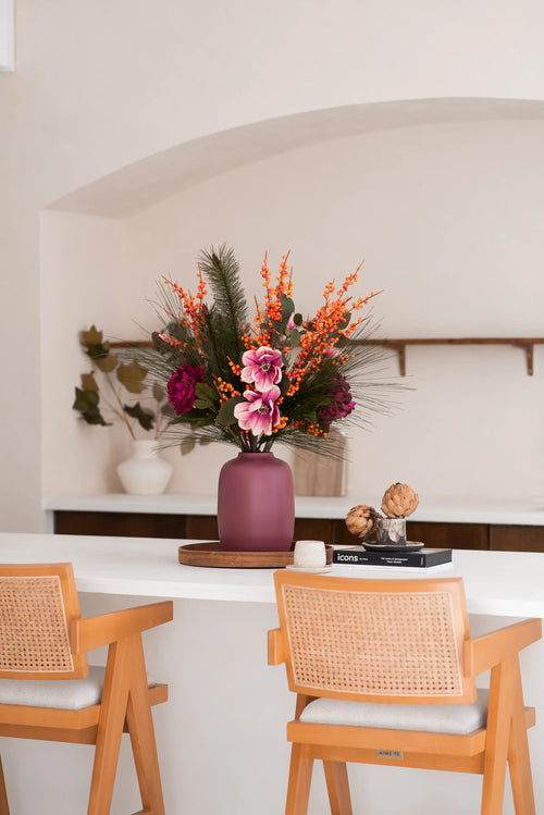 Un vaso color malva con il bouquet di fiori artificiali in seta Cozy Winter - perfetto per la decorazione autunno-inverno - si trova su un bancone bianco tra sedie di legno, con libri, carciofi, scaffali e un vaso bianco con rami nelle vicinanze.