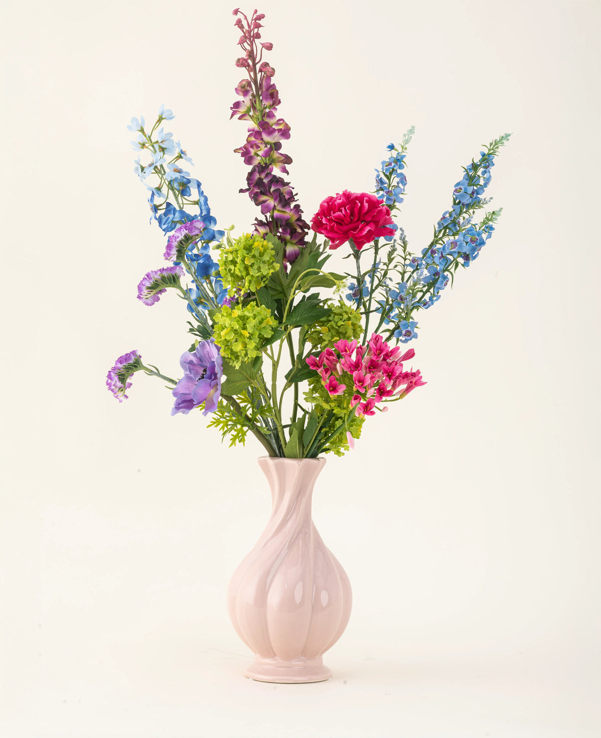 Disposto in un vaso di ceramica rosa, il bouquet di fiori artificiali in seta Blossom Breeze presenta vivaci fiori blu, viola, rossi e verdi: un bouquet floreale che non richiede manutenzione su uno sfondo chiaro e semplice.
