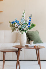 Een Zijden kunstbloemen boeket Cool Breeze van blauwe en witte bloemen staat in een witte vaas op een ronde houten salontafel met een kopje, kaarsen en een boek. Daarachter staat een witte bank met groene en beige kussens.