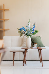 Een moderne woonkamer met een witte bank, een groen kussen en een klein houten tafeltje met daarop een Zijden kunstbloemen boeket Cool Breeze in een witte vaas, plus een koffiekopje en boeken, tegen een beige muur met een minimalistische plank.