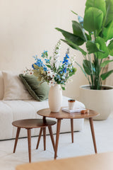 In een gezellige woonkamer staan twee houten nesttafels met een witte vaas met daarin het Zijden kunstbloemen boeket Cool Breeze, plus een kopje, boek en kaars. Een witte bank met groene kussens en een grote plant in pot maken het luchtige decor compleet.