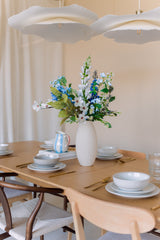 Een houten eettafel gedekt met borden, schalen, goudkleurig keukengerei en een witte vaas gevuld met het Zijden kunstbloemen boeket Cool Breeze. Zachte verlichting en moderne witte hanglampen erboven creëren een uitnodigende sfeer.