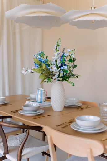 Een houten eettafel gedekt met borden, schalen, goudkleurig keukengerei en een witte vaas gevuld met het Zijden kunstbloemen boeket Cool Breeze. Zachte verlichting en moderne witte hanglampen erboven creëren een uitnodigende sfeer.