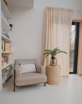 Un accogliente angolo lettura con una poltrona beige, un tavolino rotondo in vimini con sopra la pianta artificiale Baby Cycas Palm da 60 cm, librerie e tende a rete che pendono da una porta a vetri che si apre sul giardino.