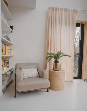 Un accogliente angolo lettura con una poltrona beige, un tavolino rotondo in vimini con sopra la pianta artificiale Baby Cycas Palm da 60 cm, librerie e tende a rete che pendono da una porta a vetri che si apre sul giardino.