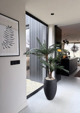 Een modern, minimalistisch interieur met een Kunstpalm Cycas Classic 135 cm in een stijlvolle sierpot bij een raam van vloer tot plafond, een varen-kunstwerk aan de muur en een strakke zwarte keuken met hanglampen op de achtergrond.