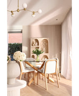 Een moderne eethoek heeft een ronde glazen tafel, houten stoelen met witte kussens en het Zijden kunstboeket Tulpen in vaas Wit als middelpunt. Decoratieve planken met vazen en transparante gordijnen maken de beige ruimte compleet.