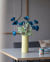 Een Kunstbloem Poppy van 73 cm in donkerblauw staat in een gele vaas op een tafel, vergezeld van een klein, kleurrijk gestreept kopje, in een moderne, zacht verlichte kamer met een minimalistische inrichting en neutrale tinten.