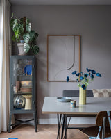 De moderne eethoek is voorzien van een grijze glazen vitrinekast waarin serviesgoed en planten zijn uitgestald, minimalistische wanddecoraties en een tafel waarop de Kunstbloem Poppy 73 cm donkerblauw in een gele vaas staat. De ruimte wordt gecompleteerd door een eclectische mix van stoelen, allemaal tegen een achtergrond van grijze muren en een houten vloer.