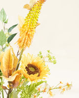 Het Zijden kunstbloemen boeket Amber Light heeft een gele zonnebloem, een lange geeloranje stekelige bloem, groene bladeren en kleine witte bloemen tegen een lichtcrème achtergrond met een amberkleurige gloed.