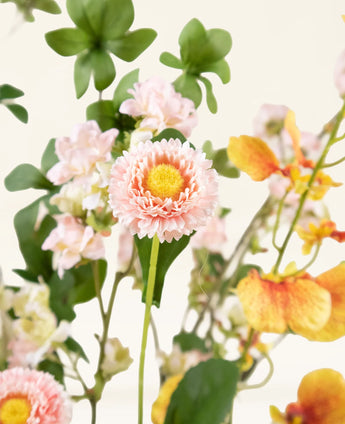 Een close-up van het Zijden kunstbloemen boeket Blossom Harmony, met een roze margrietachtige bloem met een geel hart, omringd door lichtroze bloesems en oranje-gele orchideeën, tegen een lichte achtergrond.