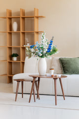 Een moderne woonkamer met een bank met witte structuur, een groen kussen, houten salontafels en een witte vaas met het Zijden kunstbloemen boeket Cool Breeze. Een houten plank met minimale decoratie maakt deze serene omgeving compleet.