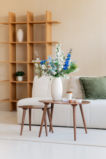 Een moderne woonkamer met een bank met witte structuur, een groen kussen, houten salontafels en een witte vaas met het Zijden kunstbloemen boeket Cool Breeze. Een houten plank met minimale decoratie maakt deze serene omgeving compleet.