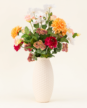 Het Zijden kunstbloemen boeket The Wild Bunch, met rode rozen, Real Touch Dahlia, roze bloemen en groene bladeren in een vaas met witte structuur, wordt getoond tegen een lichte achtergrond.