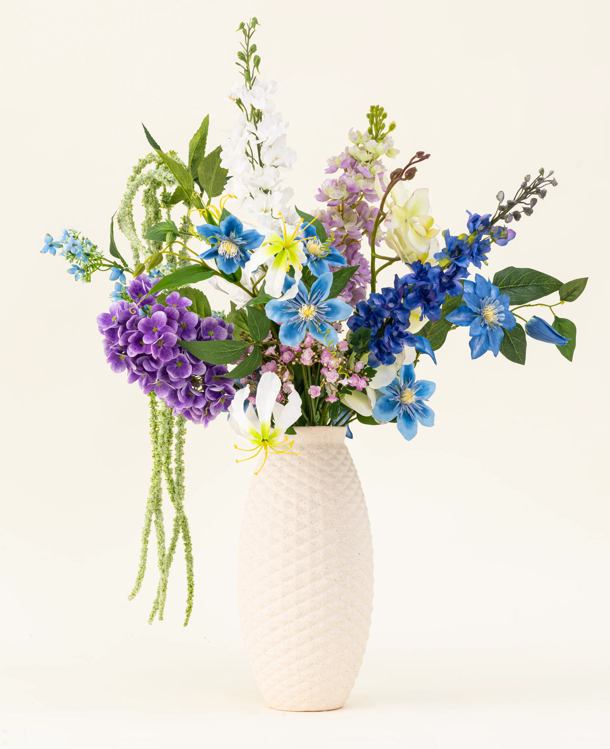Un vaso strutturato bianco contiene il bouquet di fiori artificiali in seta Midnight Valley, caratterizzato da vivaci fiori Real Touch blu, viola e gialli con diverse foglie verdi su uno sfondo chiaro.