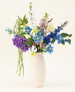 Een vaas met witte structuur bevat het Zijden kunstbloemen boeket Midnight Valley, met levendige blauwe, paarse en gele Real Touch bloemen met verschillende groene bladeren tegen een lichte achtergrond.