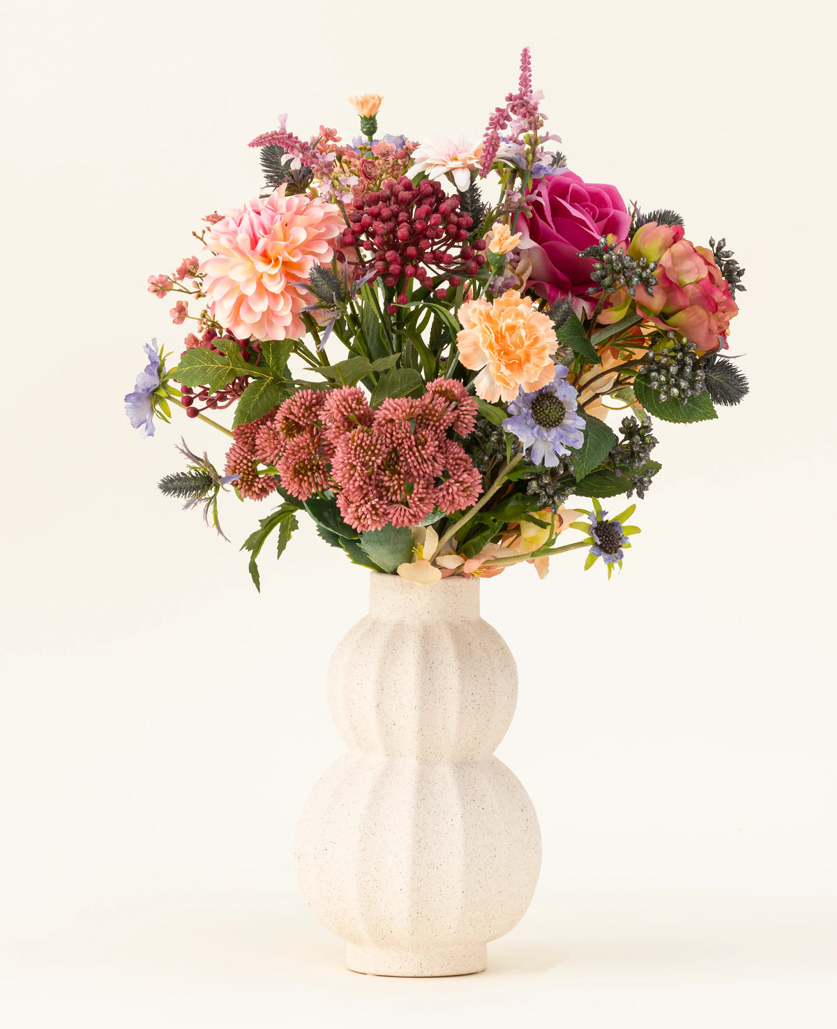 In un vaso di ceramica bianca e ricurva si trova il bouquet di fiori artificiali in seta Pretty Pastel - un mix colorato di fiori sostenibili come rose rosa, dalie, fiori viola e arancioni e vegetazione - splendidamente disposti su uno sfondo chiaro.
