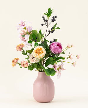 Het Zijden kunstbloemen boeket Touch of Cloudlight, gepresenteerd in een roze keramieken vaas, bevat roze pioenrozen, gele rozen, kleine bloesems, groen en takken met donkere bessen tegen een lichte achtergrond.