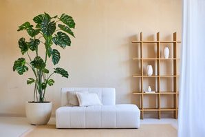 Soggiorno minimalista con una pianta artificiale di Monstera da 240 cm in un vaso, un moderno divano bianco con cuscino e un armadio aperto in legno con vasi bianchi contro una parete beige. Luce naturale da destra.