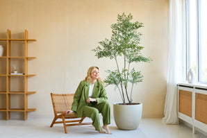 Een vrouw in een groen pak glimlacht terwijl ze op een houten stoel naast de Kunstplant Ruscus 180 cm zit in een lichte, moderne kamer met beige muren, grote ramen en een minimalistische inrichting.