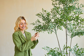 Una donna sorridente con un blazer verde è in piedi all'interno e tocca delicatamente le foglie di una pianta artificiale di Ruscus di 150 cm con rami sottili e fogliame verde lussureggiante, che si stagliano su uno sfondo semplice e di colore chiaro.