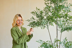 Una donna sorridente con un blazer verde è in piedi all'interno e tocca delicatamente le foglie di una pianta artificiale di Ruscus di 150 cm con rami sottili e fogliame verde lussureggiante, che si stagliano su uno sfondo semplice e di colore chiaro.