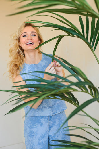 Een glimlachende vrouw met lang blond haar, in een lichtblauwe mouwloze top en bijpassende broek, staat binnen achter de Kunstplant Kentia Palm 180 cm en raakt speels een van de groene bladeren aan.