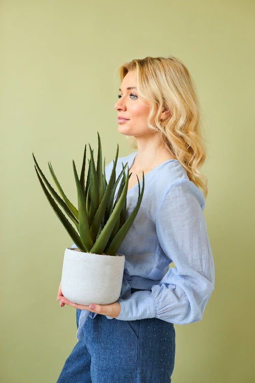Una donna con lunghi capelli biondi in una camicetta azzurra e pantaloni blu tiene la pianta artificiale Aloe Vera da 43 cm in un vaso su uno sfondo verde chiaro.