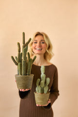Een glimlachende vrouw in een bruine jurk houdt twee Kunst Cactus Cereus 35 cm planten in pot, een grote en een kleine, tegen een effen beige achtergrond.