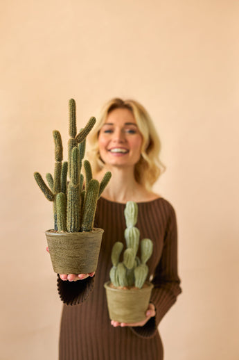 Een glimlachende vrouw in een bruine jurk houdt twee Kunst Cactus Cereus 35 cm planten in pot, een grote en een kleine, tegen een effen beige achtergrond.