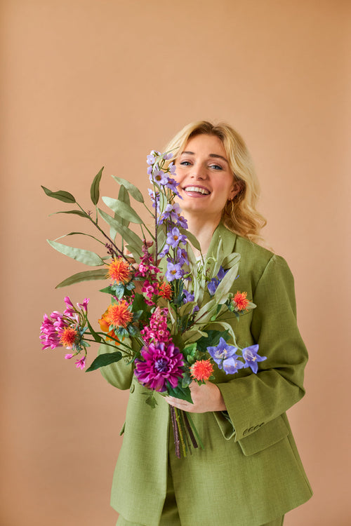 Una persona bionda sorridente in un abito verde tiene il vivace bouquet di fiori artificiali in seta Svenski colorata su uno sfondo marrone chiaro.