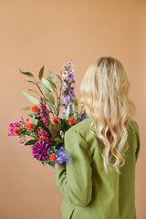 Una persona con lunghi capelli biondi ondulati e una giacca verde è vista da dietro, mentre tiene in mano il bouquet di fiori artificiali in seta Colourful Svenski su uno sfondo beige semplice.
