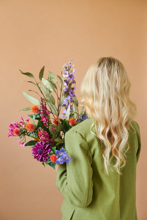 Una persona con lunghi capelli biondi ondulati e una giacca verde è vista da dietro, mentre tiene in mano il bouquet di fiori artificiali in seta Colourful Svenski su uno sfondo beige semplice.