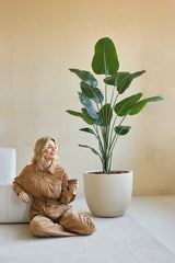 Una donna con una tuta marrone siede sorridente sul pavimento con una tazza di caffè accanto, accanto alla pianta artificiale Strelitzia 180 cm, un vero tocco in una stanza minimalista con pareti beige.