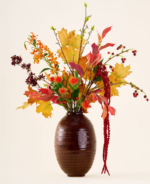 In un vaso marrone c'è il bouquet 'Rustic Glow' di fiori artificiali di seta, con foglie gialle, arancioni e rosse, bacche rosse, fiori d'arancio e lunghi amaranto rosso pendenti su uno sfondo neutro.
