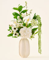 In un vaso di ceramica bianca si trova il bouquet "Green Elegance" di fiori artificiali di seta, una composizione di ortensie bianche, calle, anemoni, foglie verdi e vegetazione pendente su uno sfondo chiaro e semplice.