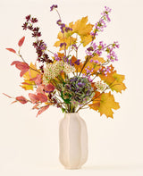 Il bouquet di fiori artificiali di seta "Wild Amethyst", con foglie autunnali, fiori viola e bianchi e ortensie verdi, è in un vaso di ceramica bianca su uno sfondo semplice e chiaro.