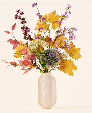 Il bouquet di fiori artificiali di seta "Wild Amethyst", con foglie autunnali, fiori viola e bianchi e ortensie verdi, è in un vaso di ceramica bianca su uno sfondo semplice e chiaro.