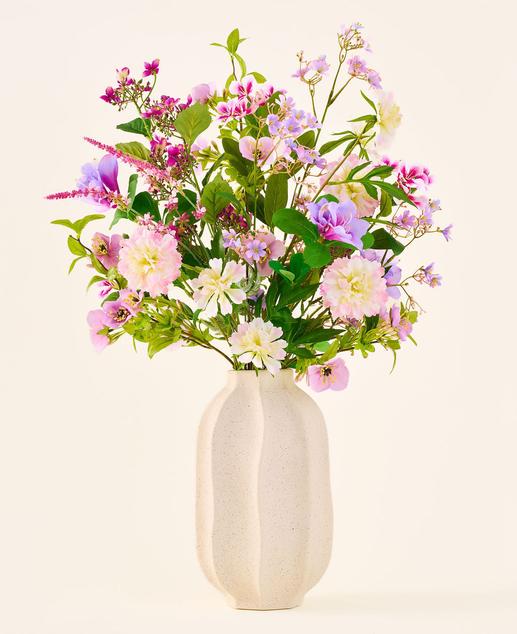 Un vaso in ceramica bianca contiene un bouquet colorato di fiori rosa, viola e bianchi della collezione "Bouquet di fiori artificiali in seta Royal Bloom", accentuato da foglie verdi e presentato su uno sfondo semplice e chiaro.