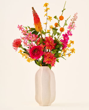 In un vaso di ceramica bianca si trova il bouquet di fiori artificiali di seta "Summer Blush", composto da fiori rosa, gialli e arancioni con foglie verdi, su uno sfondo chiaro e semplice.