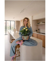 Een vrouw met krullend blond haar, gekleed in spijkerkleding en roze sneakers, zit in kleermakerszit op een keukeneiland in een moderne, zonovergoten keuken, met de Kunstplant Gardenia UV 39 cm in haar handen.